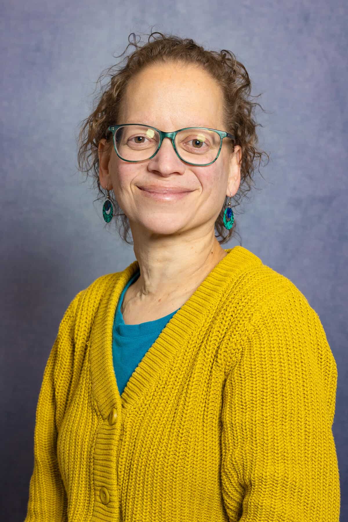 Headshot of a white woman with short hair wearing glasses, earrings and a mustard yellow cardigan